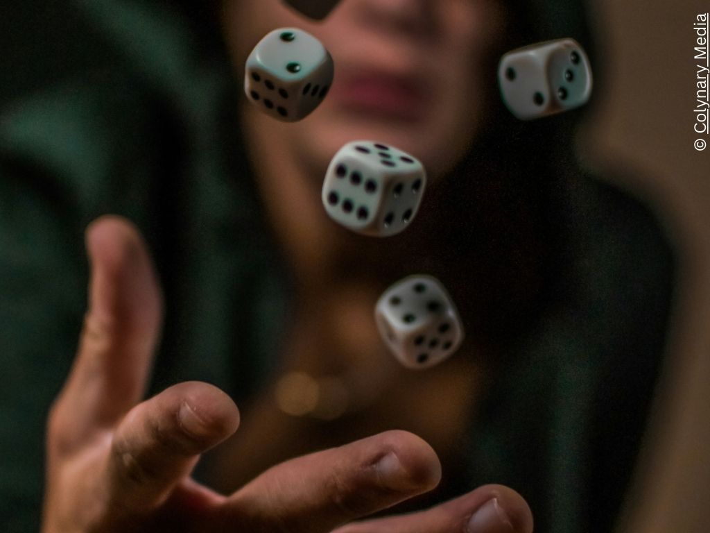 A hand throws five dice in the air. There is a hooded face in the background.