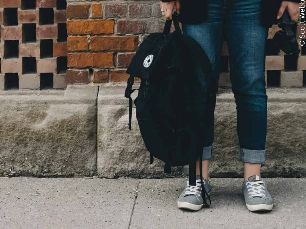 A photo of the curb. We can see the legs of a girl in jeans and trainers. She has a backpack at her feet.