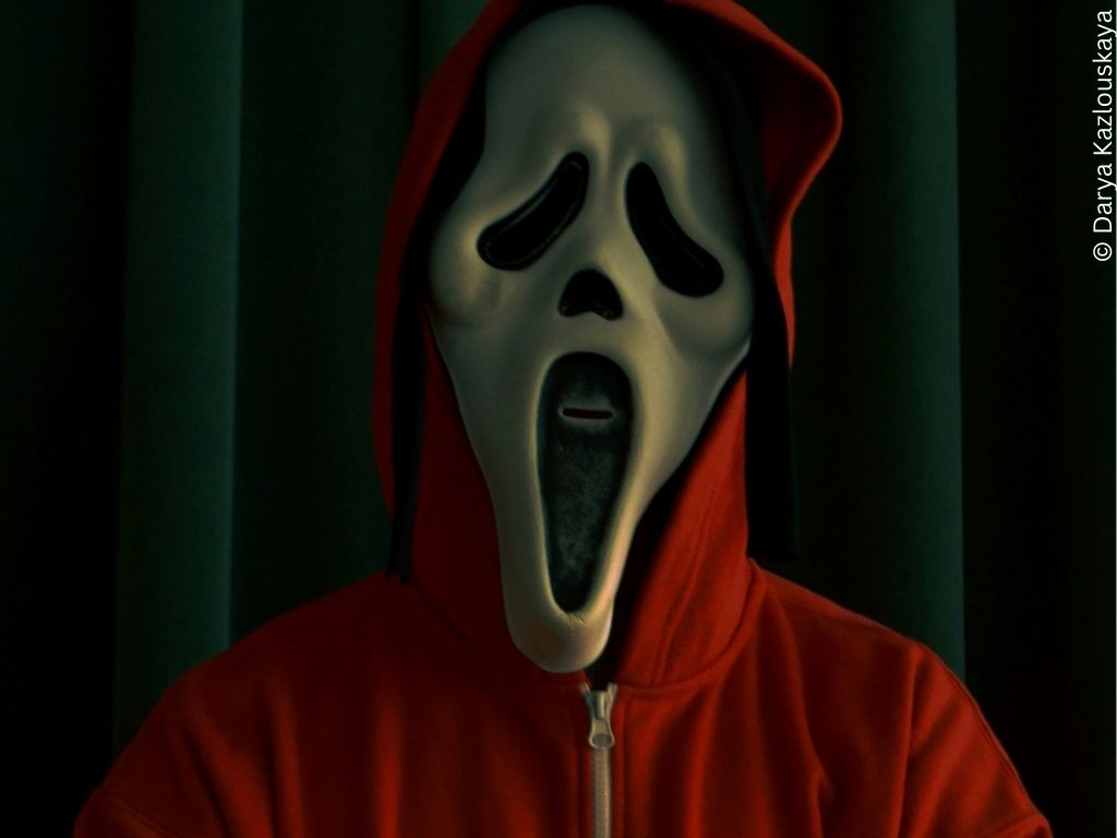A person wearing the white mask from the film 'Scream' looks towards the camera.