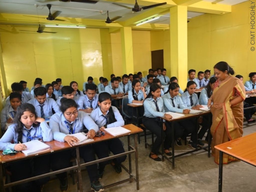 A group of Indian girls sit at desks in a school. A teacher stands at the front of the class.