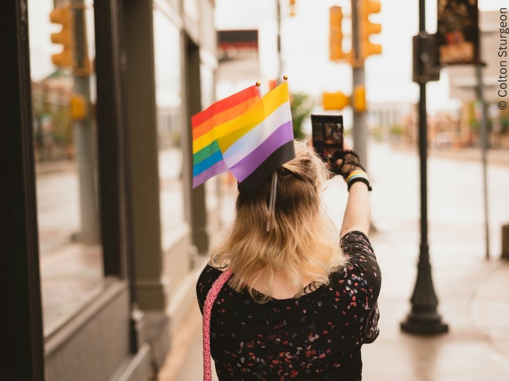 The back view of a young person in the street. They carry two LGBT+ flags.