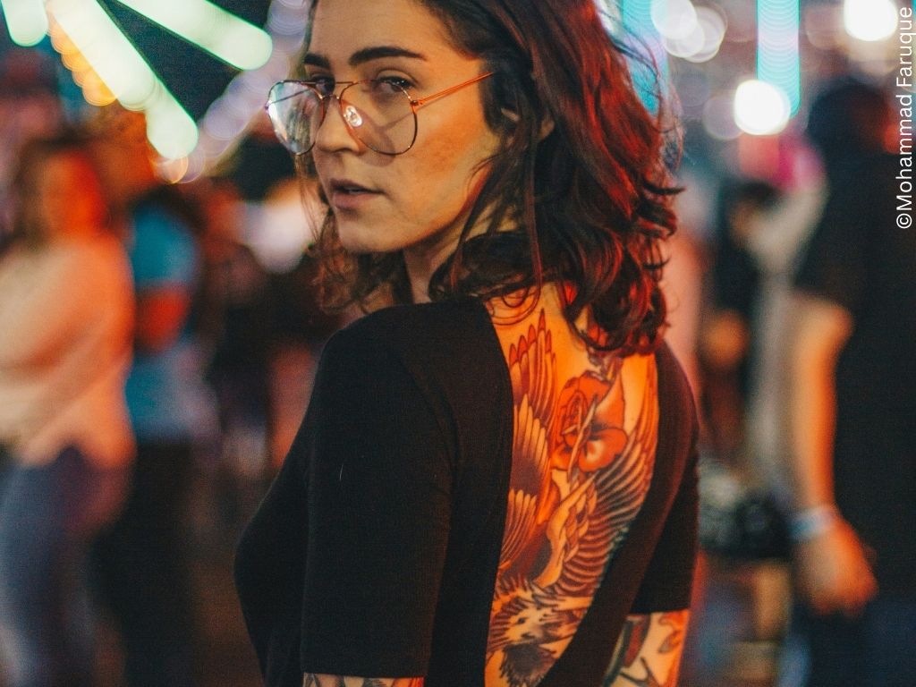 A young woman with glasses and tattoos looks back over her shoulder. She may be at a fair.