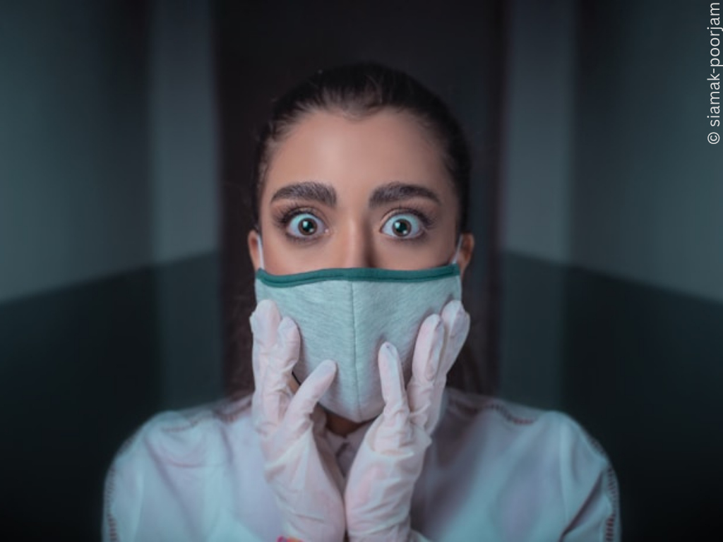 A young brown woman looks alarmed. She wears a medical mask and gloves.