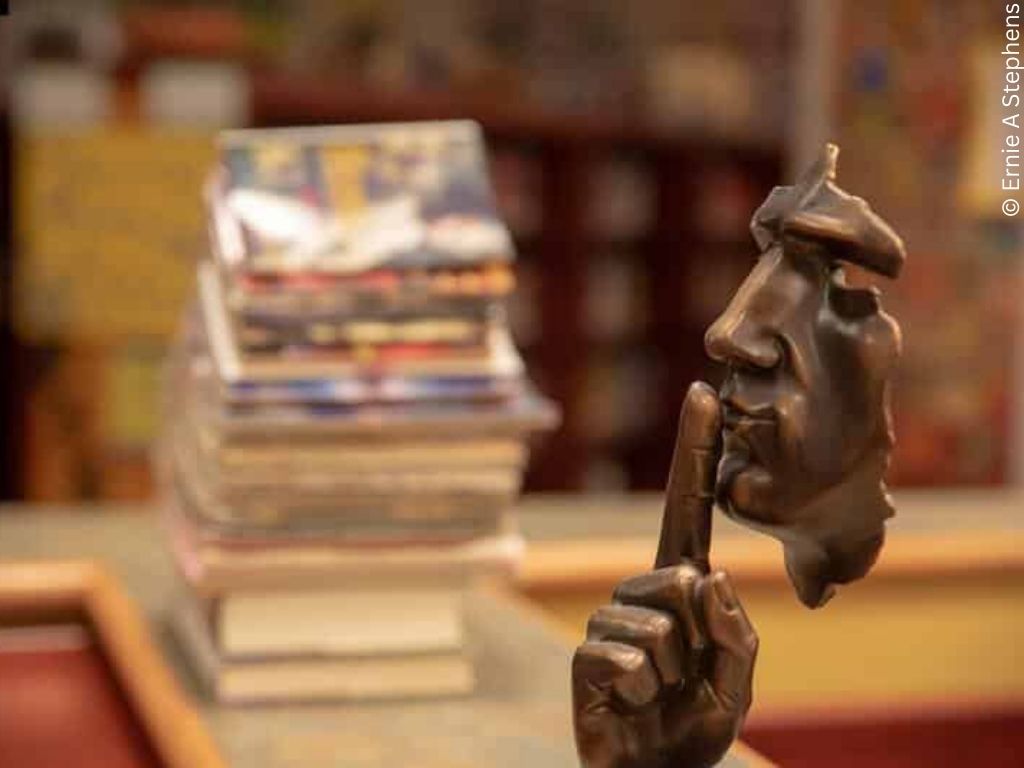 A brass statuette of a face holds up a finger for silence. In the background is a stack of books.