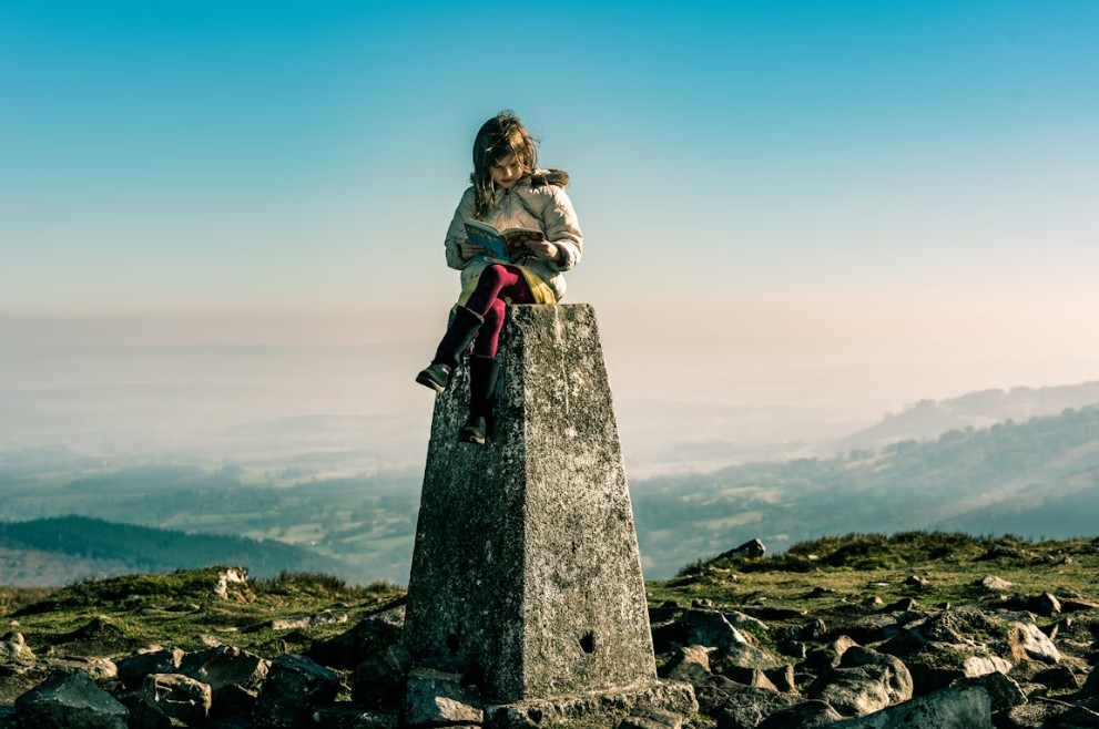 A girl sits on top of a rock. She is reading. Behind her is a mountainous landscape